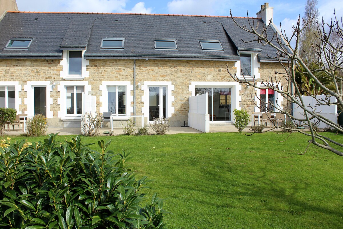 A two-story stone house is set in a landscaped garden, featuring multiple large windows. Sunny outdoor seating is provided on a patio area, while green grass and shrubs frame the property. The structure displays a blend of classic stonework and modern design.