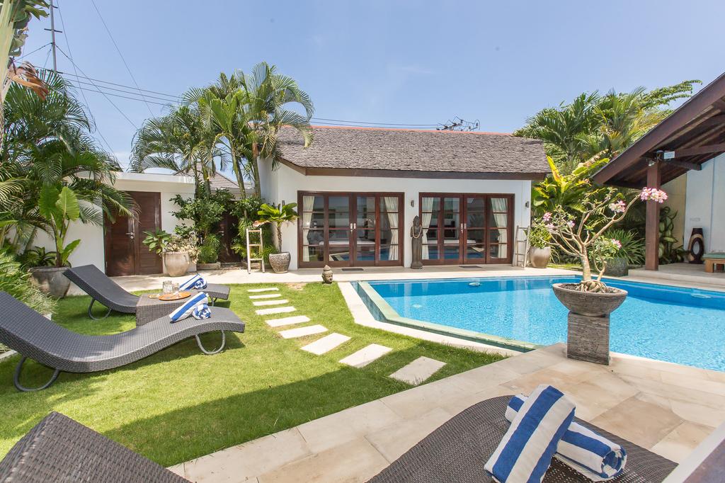 The outdoor area features a large pool surrounded by well-maintained grass and tropical plants. Two loungers sit near the pool, positioned for relaxation. A pathway of stones leads toward the building, which has expansive glass doors and a traditional roof.