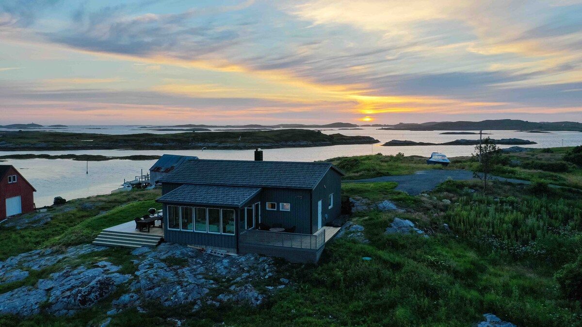 A coastal cabin is set against a scenic sunset, with vibrant orange and purple hues reflecting over calm waters. The structure is surrounded by greenery and rocky terrain, with a deck that invites outdoor relaxation and provides panoramic views of the serene landscape.