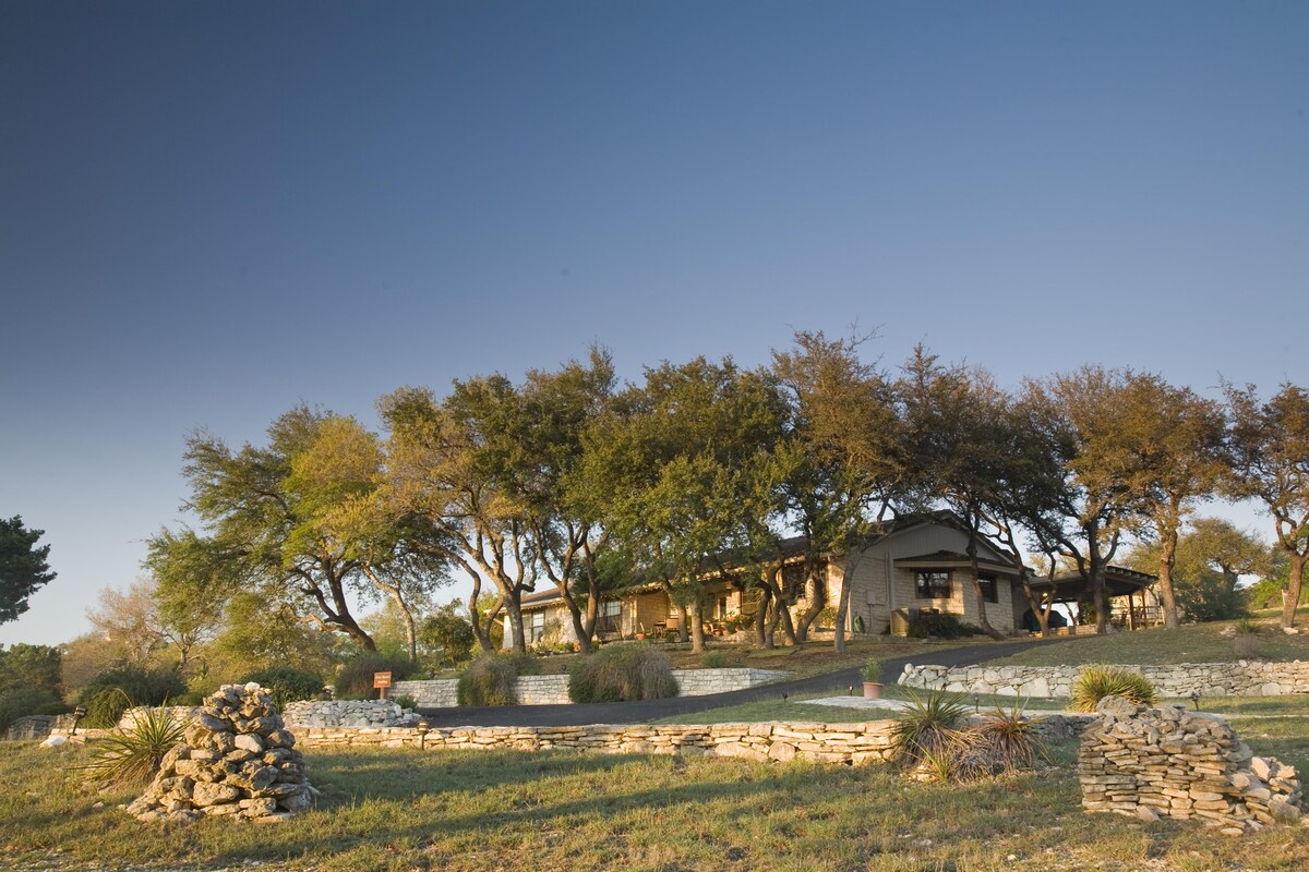 The cottage is set amid a natural landscape, surrounded by trees and grassy areas. A stone pathway leads to the structure, which features a warm-toned exterior. Rock formations and landscaping elements add a rustic charm to the setting, enhancing the tranquil ambiance of the hill country.
