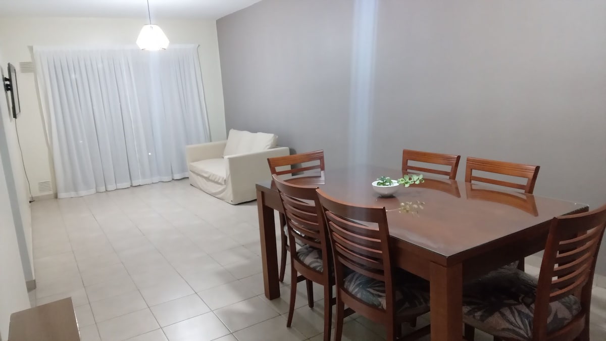A dining area features a wooden table surrounded by six chairs, providing ample seating. A soft sofa is positioned beside sheer white curtains, allowing natural light to filter into the space. Neutral tones and tiled flooring enhance the room's airy feel.
