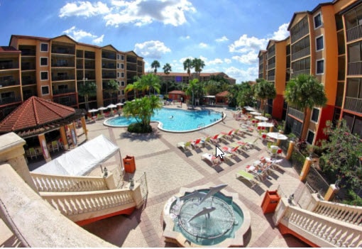 An expansive pool area is visible, surrounded by lounge chairs under bright umbrellas. A large swimming pool takes center stage, with a smaller fountain feature nearby. Lush palm trees frame the space, and colorful buildings provide a vibrant backdrop.