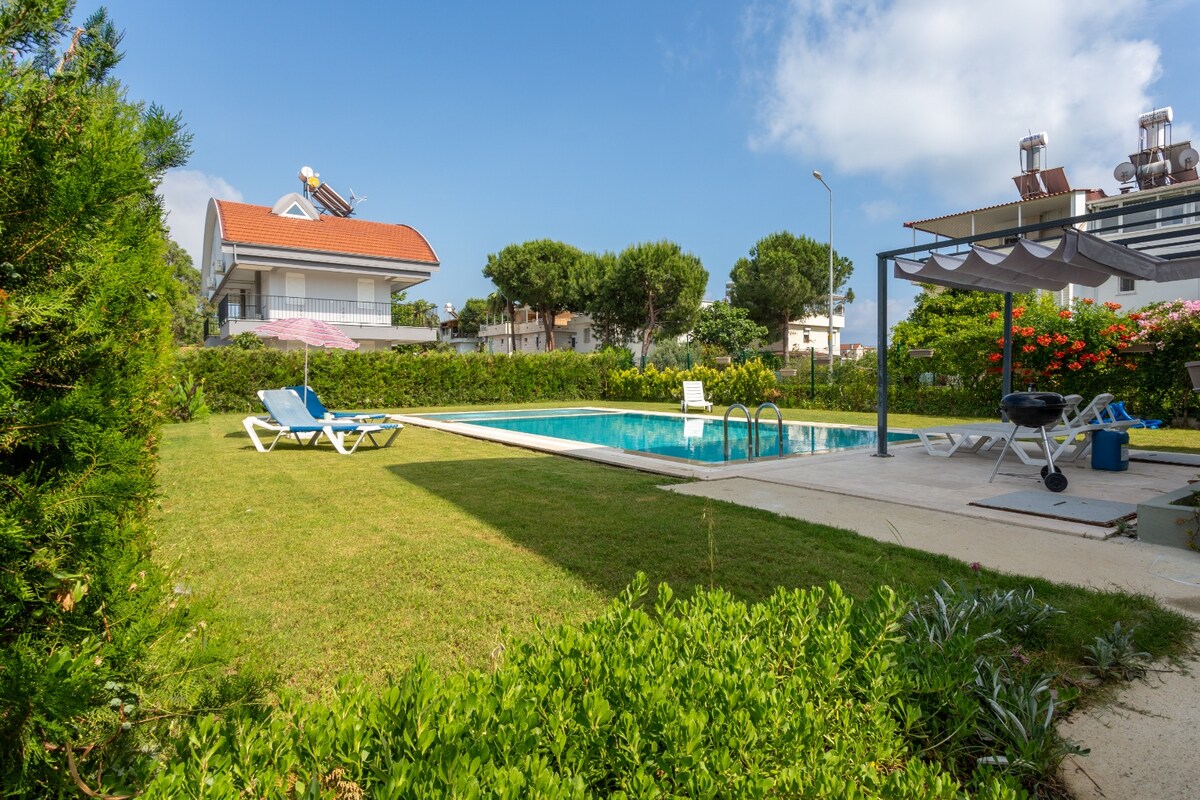 The image showcases a spacious grass-covered garden surrounding a shared pool. Sun loungers are positioned near the water. Lush greenery and flowering shrubs create a serene atmosphere, while a shaded seating area is visible on the right, perfect for relaxation.