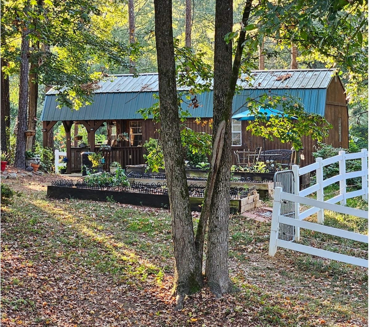 A rustic wooden structure with a green metal roof is visible, surrounded by trees that provide dappled sunlight. Outdoor seating is seen on the porch, overlooking a landscaped yard with garden beds and a white fence in the foreground.