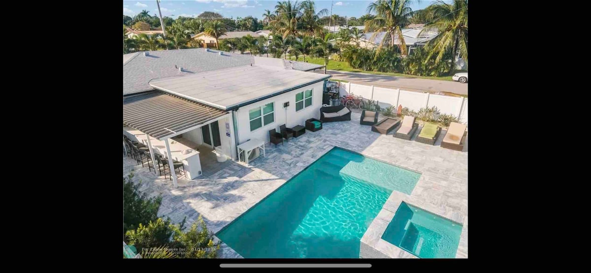 A spacious outdoor area features a heated saltwater pool alongside a separate jacuzzi. Modern lounge furniture is arranged around the pool deck, providing ample seating. Well-maintained landscaping surrounds the space, with palm trees in the background, creating a relaxed atmosphere.