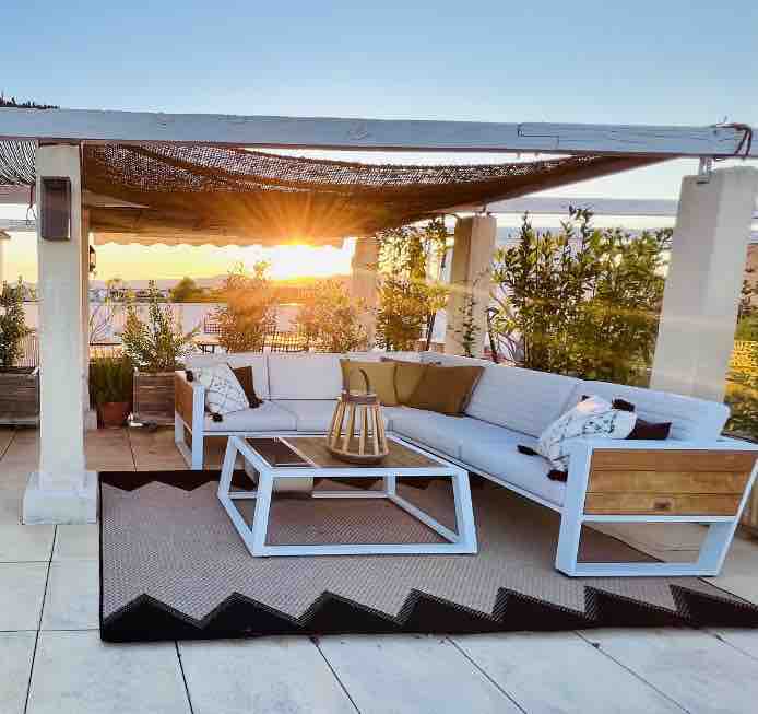 A spacious rooftop area is shown, featuring a comfortable seating arrangement with white cushions on a sectional sofa. A coffee table sits in front, placed on a patterned rug. The setting is enhanced by greenery surrounding the area, with a sunset visible in the background.