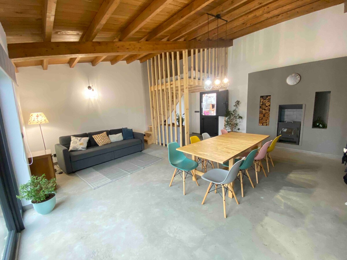 A spacious living and dining area is characterized by vaulted wooden ceilings and large windows allowing natural light. A wooden dining table surrounded by colorful chairs offers ample seating. A comfortable sofa is positioned nearby, complemented by a decorative lamp and a potted plant.