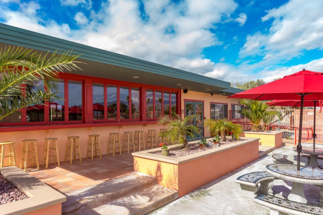 An outdoor patio area features several tall stools arranged along a counter, surrounded by lush palm plants. Bright red umbrellas provide shade over outdoor seating areas, complemented by smooth stone surfaces and vibrant landscaping. The building showcases large windows, inviting natural light.
