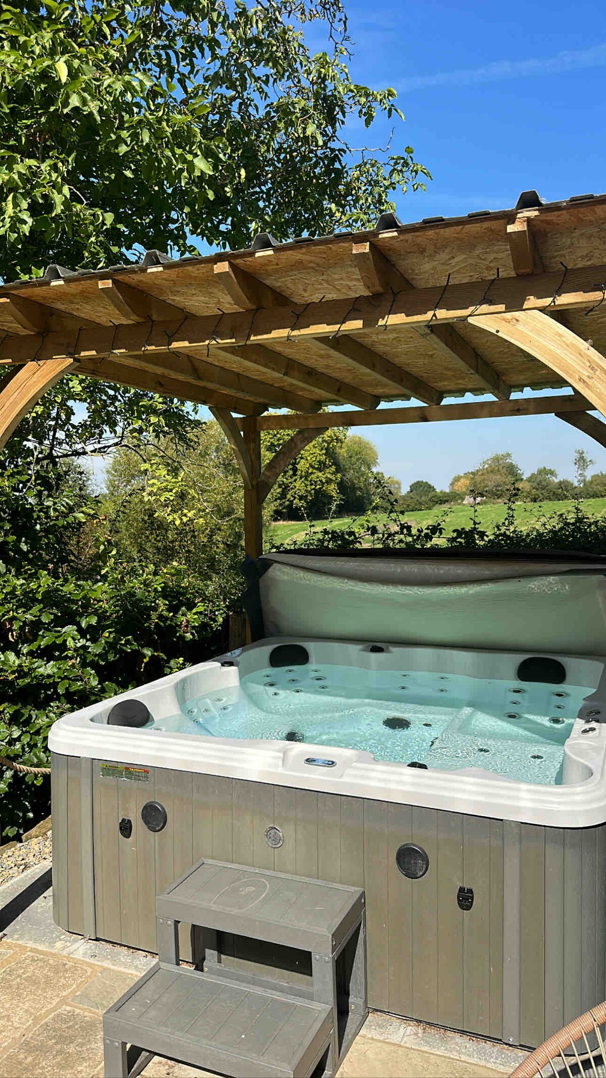 A hot tub is situated on a patio beneath a wooden gazebo, surrounded by greenery. The clear water reflects the sunlight, creating a tranquil atmosphere. A pair of steps leads up to the hot tub, enhancing accessibility.