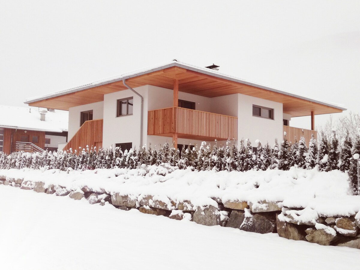 A modern building is set in a snowy landscape, showcasing a spacious balcony surrounded by wooden railings. The structure features a flat roof and large windows, with a stone wall visible in the foreground, enhancing the serene winter setting.
