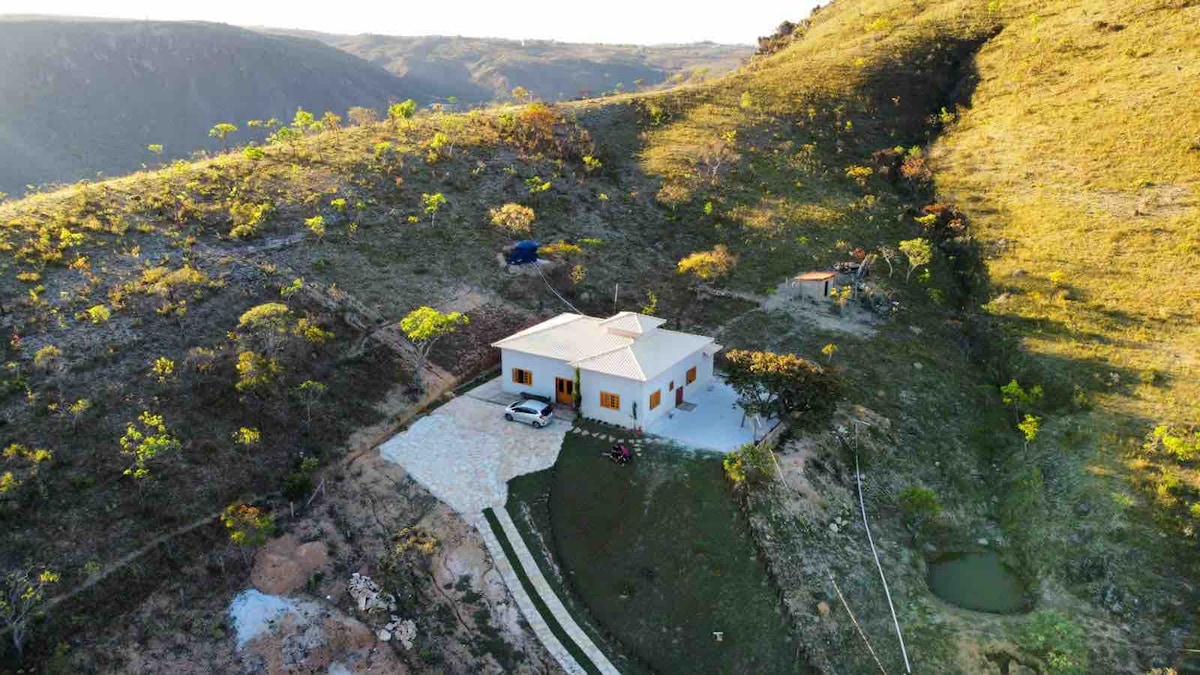 A modern hillside retreat is positioned among rolling green landscapes. The property features a light-colored exterior with multiple windows, a driveway leading to the entrance, and a garden area. Nearby, a small pond reflects the surrounding natural scenery.