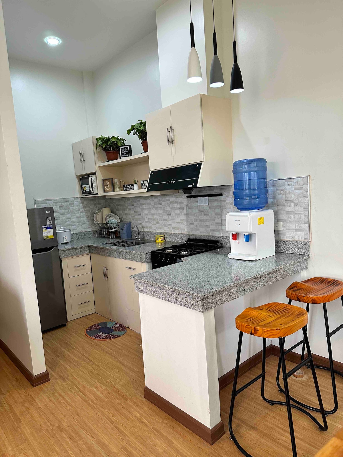 A compact kitchen features modern appliances, including a refrigerator and stove, complemented by a clean countertop. Bar stools are positioned at the counter, and a water dispenser stands nearby. Warm wood flooring enhances the overall bright and functional space.