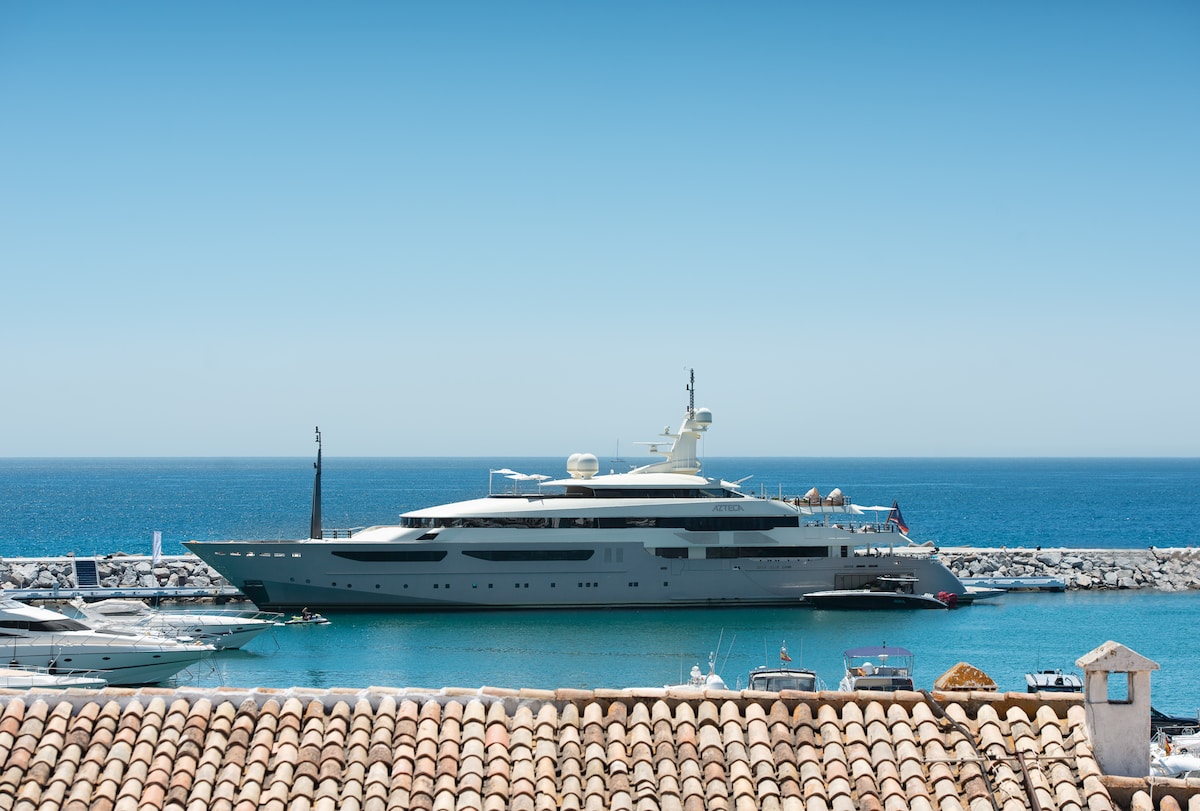 A luxurious yacht docked at a marina, featuring a sleek design and multiple levels. The vibrantly blue sea serves as a backdrop, contrasting with the clear sky above. Nearby, smaller boats can be seen, contributing to the upscale nautical atmosphere.