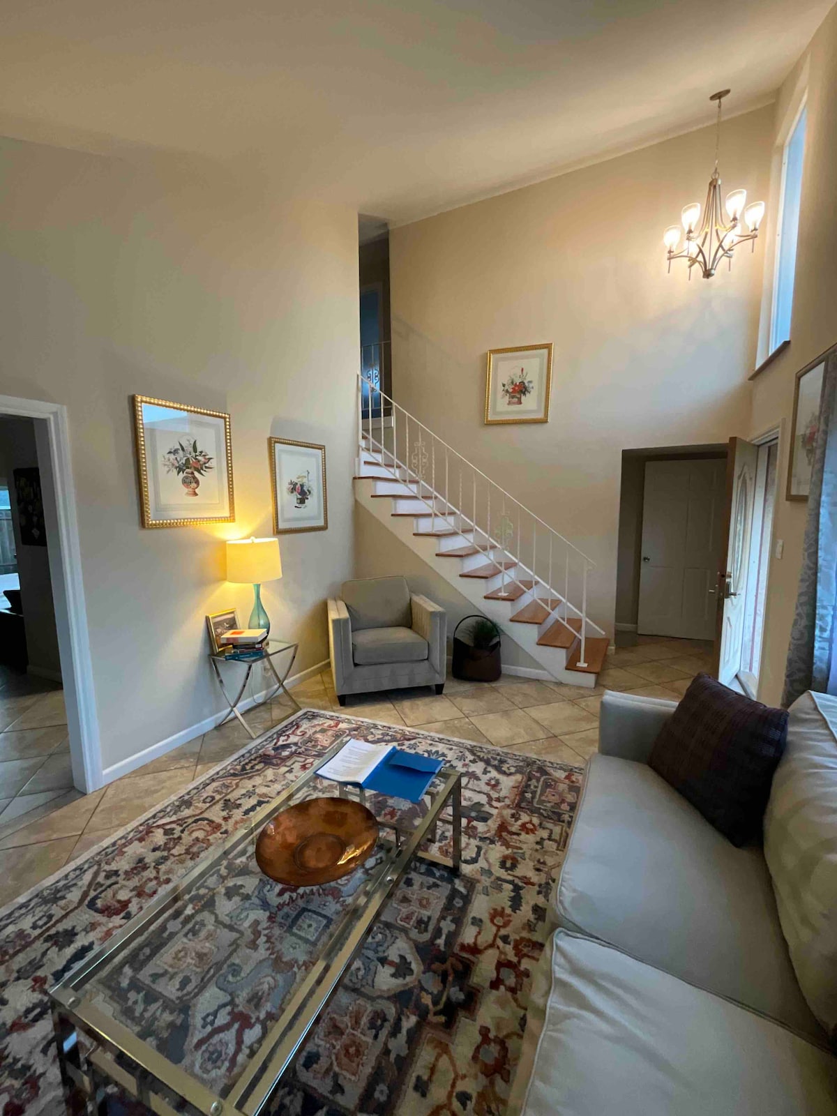 A well-lit entryway features a neutral color palette with a staircase leading to the upper floor. Elegant framed artwork hangs on the walls. A cozy seating area includes a plush armchair and a coffee table set upon a patterned area rug, enhancing the home’s inviting ambiance.