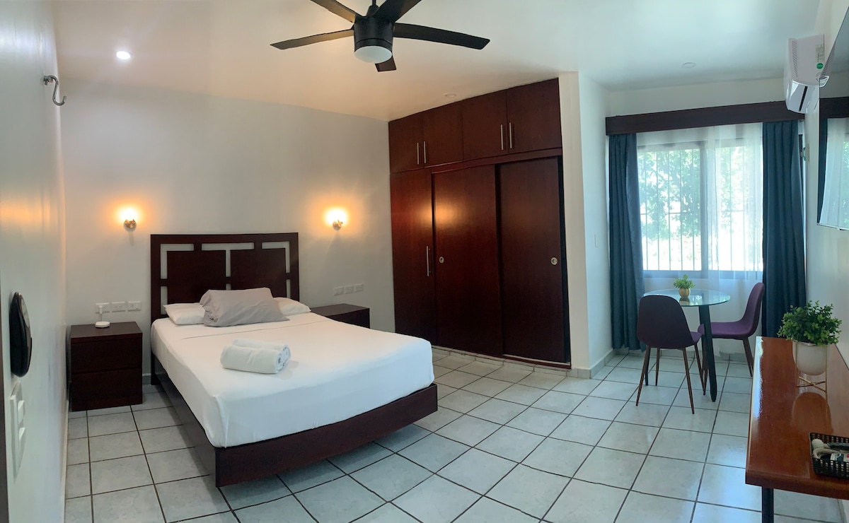 A spacious bedroom features a queen-sized bed with crisp white linens. A wooden nightstand and a large wardrobe provide ample storage. A small dining table with chairs is set by the window, allowing natural light to brighten the room. A ceiling fan is installed for comfort.