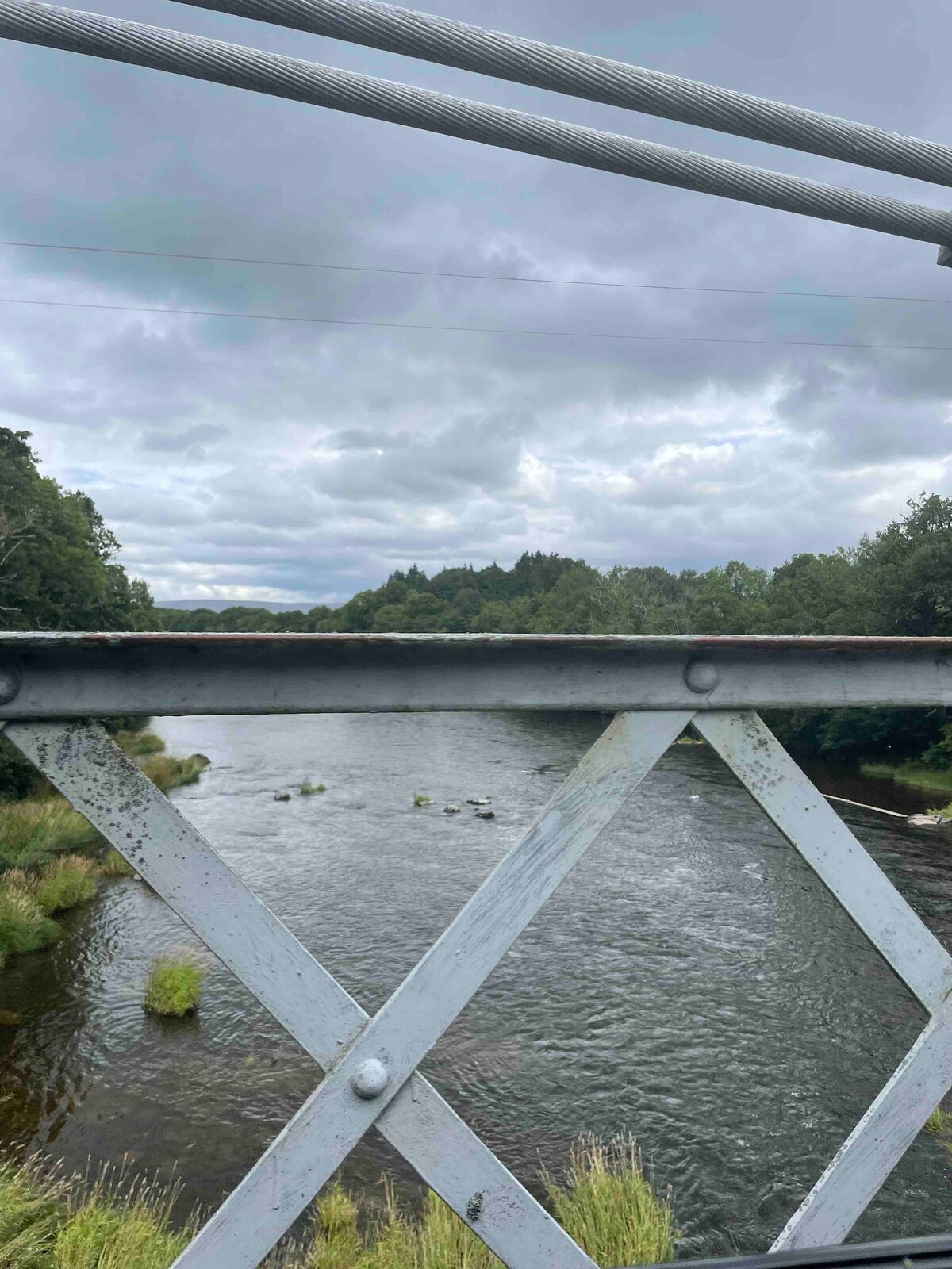 A view of a river flows underneath a metal bridge, framed by a geometric pattern of the bridge's railing. Lush greenery lines the riverbank, and rolling hills are visible in the background, all under a cloudy sky.