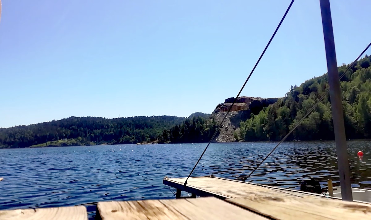 A wooden dock extends over calm water, with clear blue skies above. Lush green hills border the scene, creating a peaceful backdrop. The image captures the serene beauty of the natural surroundings, suggesting a tranquil environment for relaxation.