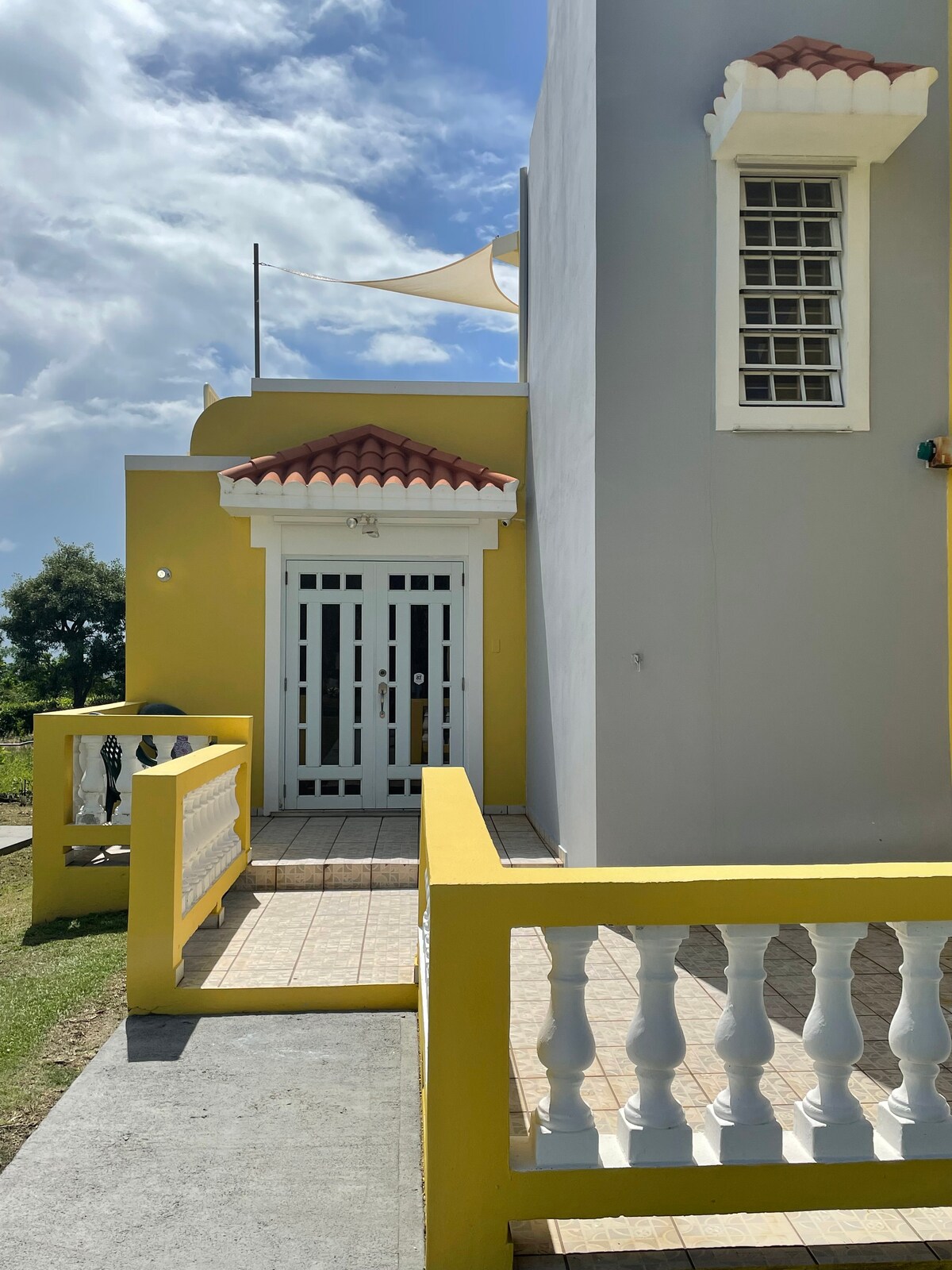 The entrance features a bright yellow façade, complemented by a covered entryway with ornate railings. Large double doors lead into the apartment, while a shaded area above offers protection from the sun. The surrounding greenery and blue sky enhance the welcoming appearance.