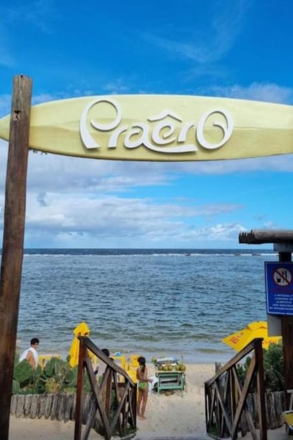 The entrance to Praero Beach is marked by a wooden sign above a sandy path leading down toward the water. Yellow umbrellas are scattered on the beach, with people enjoying the beach area. The calm water reflects a clear blue sky, enhancing the coastal atmosphere.