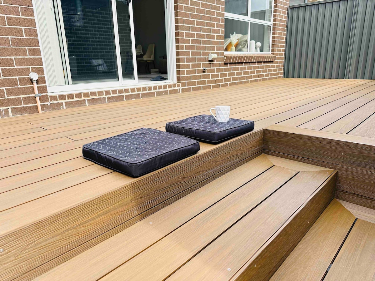 A wooden deck features two black cushions placed on the steps, with a cup resting on one. Large glass doors in the background provide a view into a well-lit interior space, emphasizing the seamless connection between outdoor and indoor areas.