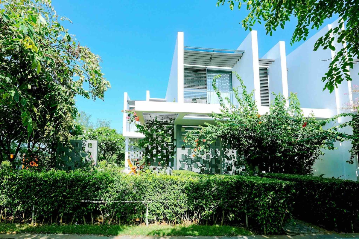 The villa's exterior is surrounded by lush greenery, with well-maintained hedges and flowering plants. The modern architecture features a blend of white walls and large overhangs, offering a covered terrace on the second floor. Clear blue skies enhance the inviting atmosphere.