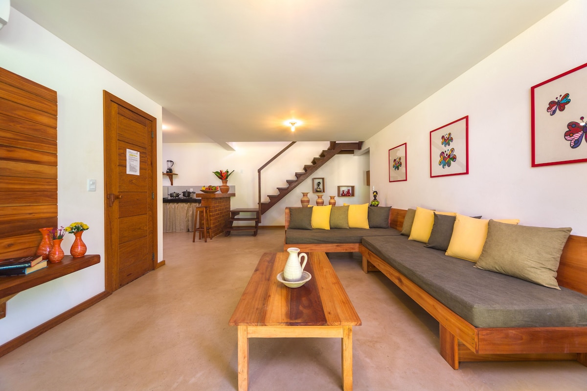 A spacious living area is highlighted by a large wooden coffee table and a comfortable L-shaped sofa adorned with colorful cushions. Framed artwork decorates the walls, and a staircase leads to the upper level, offering convenient access to additional rooms.