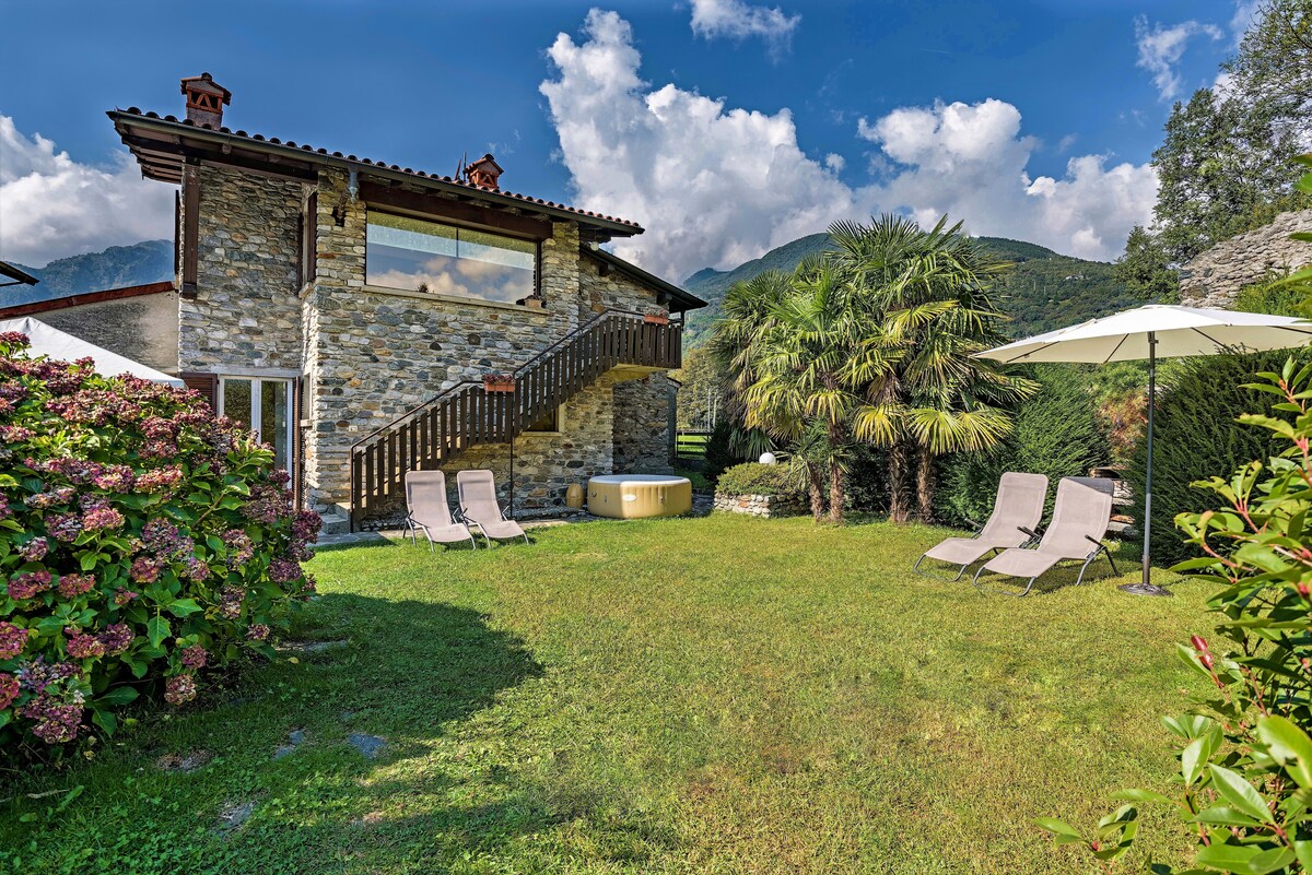 An expansive garden area features a luxurious hot tub surrounded by greenery and flowering plants. Two sun loungers are positioned on the grass, and a large sun umbrella provides shade. The stone exterior of the house is complemented by a modern staircase and mountain views in the background.