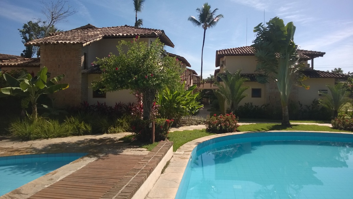 A tranquil pool area is surrounded by lush tropical gardens, featuring palm trees and vibrant greenery. Stone pathways lead toward charming, two-story buildings with terracotta roofs, adding a serene touch to the setting.