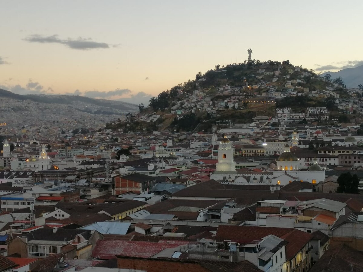 A panoramic view of Quito's historical center is captured at dusk, showcasing the rooftops of traditional architecture. The prominent hill with a statue overlooks the city, while soft twilight hues add depth to the skyline. The scene reflects the unique blend of urban and natural elements.