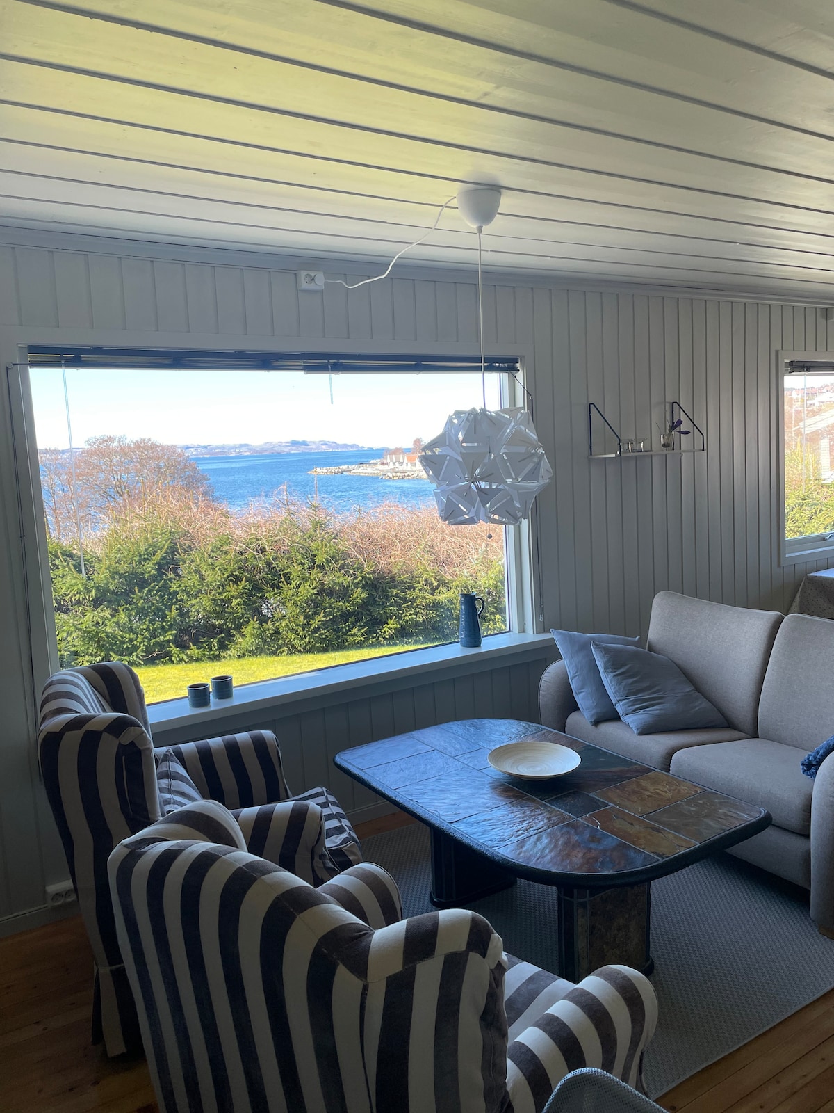 A cozy living area is presented with two striped chairs and a comfortable sofa. A wooden coffee table sits in the center. Large windows frame views of the surrounding greenery and water, allowing natural light to fill the space.
