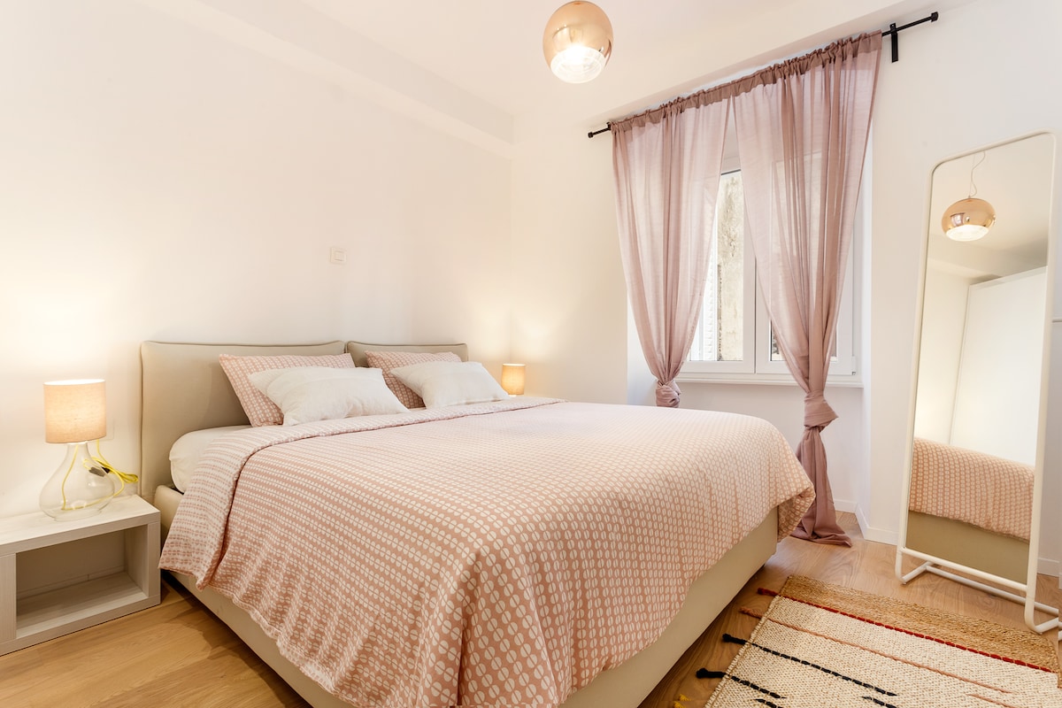 A spacious bedroom features a large bed with a light-hued, patterned blanket. Soft lighting is provided by bedside lamps and a ceiling fixture. A mirror stands beside the bed, while sheer curtains frame the window, allowing natural light to brighten the room.