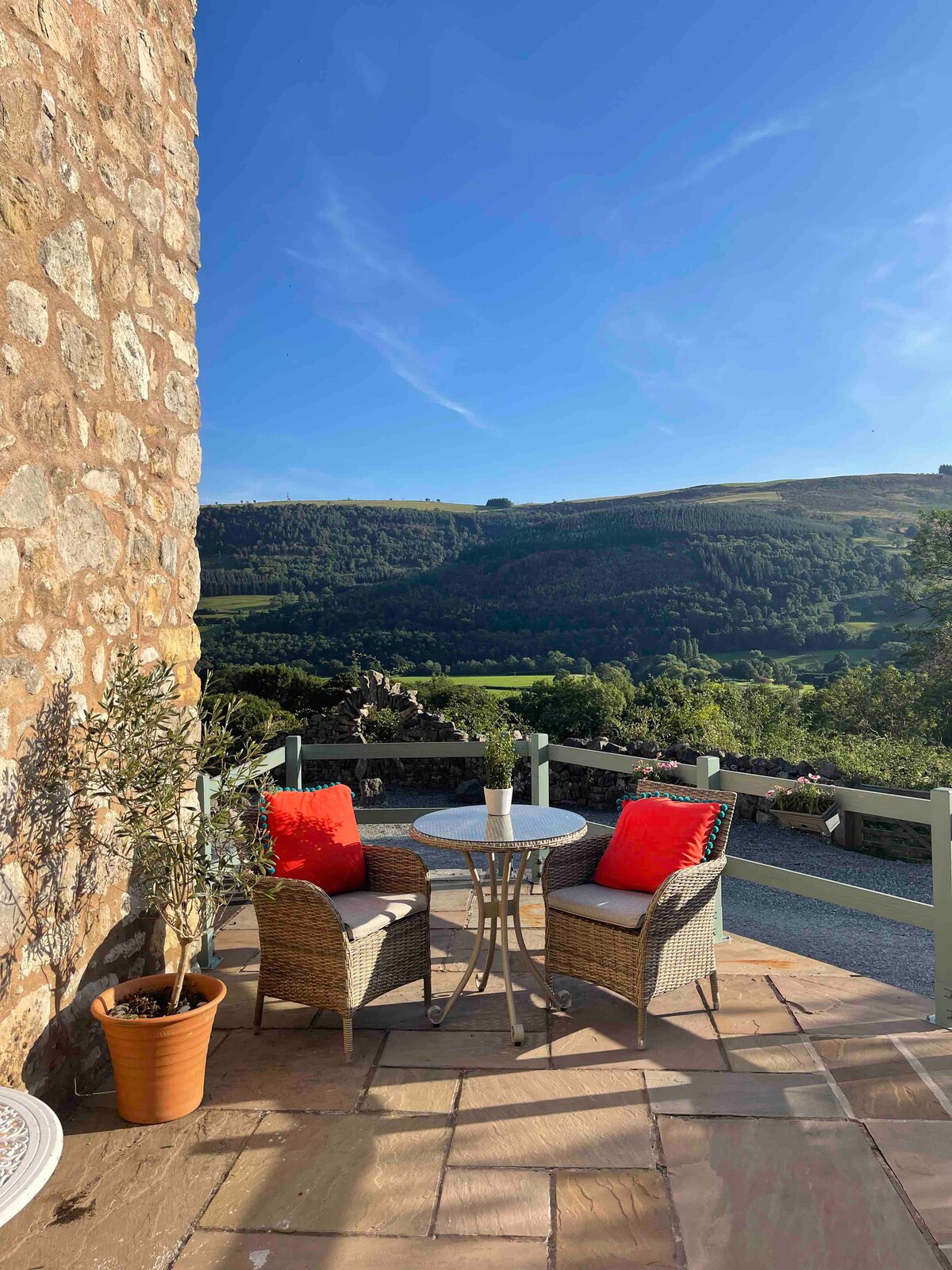 A private terrace is shown, featuring a round table and two cozy chairs adorned with bright cushions. The scenic view of the lush hills and valleys in the background complements the rustic stone wall that frames the space.