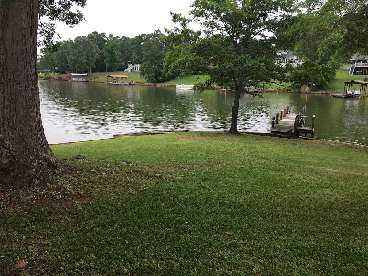 A grassy area leads to a calm lake, where trees provide shade along the shoreline. A wooden dock extends into the water, offering space for boats. In the background, gently sloping greenery is visible, adding to the serene lakeside setting.
