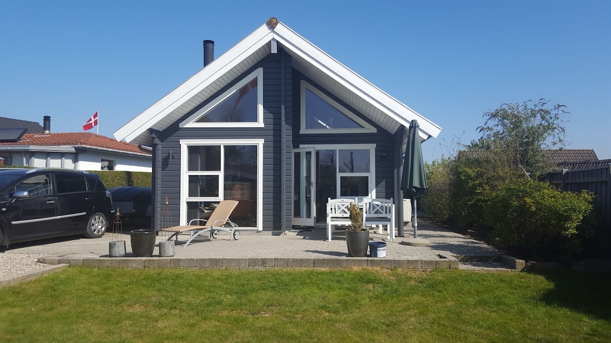 A modern blue house features large windows that invite natural light. In front, there are lounge chairs and a small table on a gravel patio surrounded by well-maintained grass. The relaxing outdoor space is framed by greenery, creating a welcoming environment.