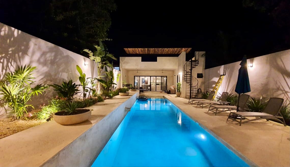 A serene outdoor area is highlighted by a lap pool, reflecting soft blue light under the night sky. Surrounding greenery and carefully arranged loungers provide a calming atmosphere. The exterior walls feature subtle lighting, enhancing the tranquil setting.