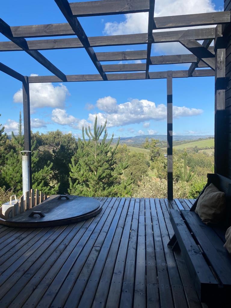 A wooden terrace is shown, featuring a circular hottub and a comfortable bench. Surrounding greenery is visible, with a scenic view overlooking undulating hills and a blue sky adorned with scattered clouds. Natural light enhances the outdoor space.