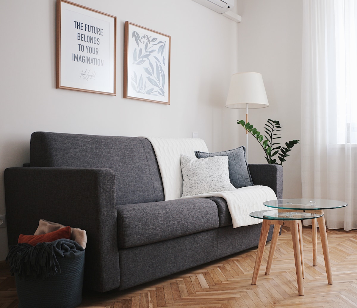 A comfortable gray sofa is positioned in the living area, adorned with a neutral throw and decorative pillows. A glass coffee table with wooden legs sits in front, while a plant adds a touch of greenery. Two framed art pieces are displayed on the wall above.