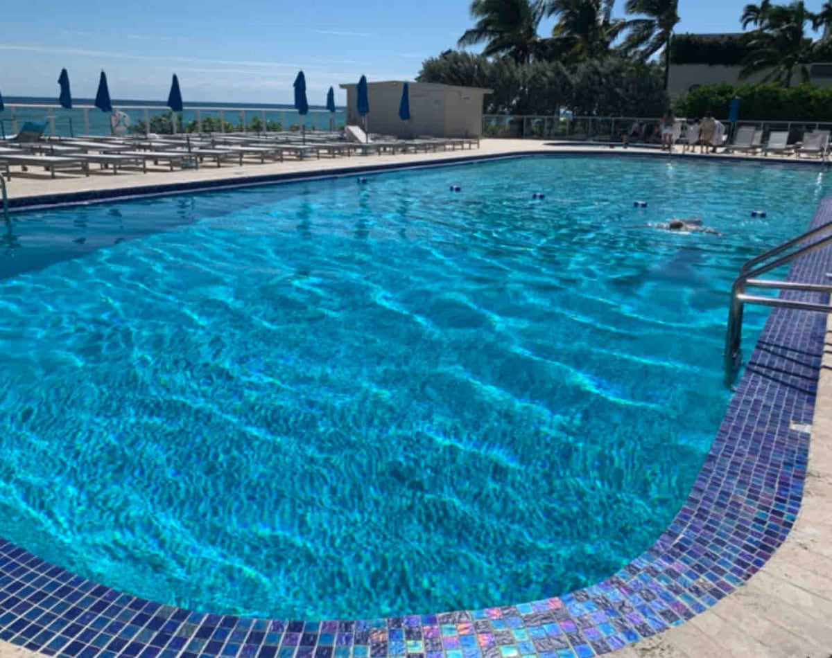 A large heated swimming pool is shown with sparkling blue water. Surrounding areas feature lounge chairs arranged under blue umbrellas. Palm trees can be seen in the background, providing a tropical ambiance.