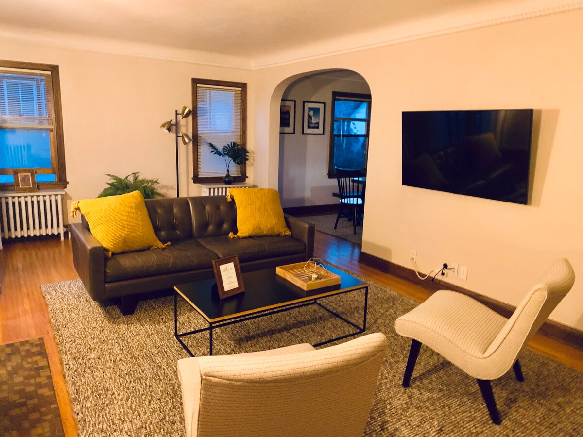 A living area is highlighted by a brown sofa adorned with yellow throw pillows. A coffee table is positioned in front, with a decorative tray. Two light-colored chairs are placed across from the sofa, and a wall-mounted television is positioned beside an arched doorway.