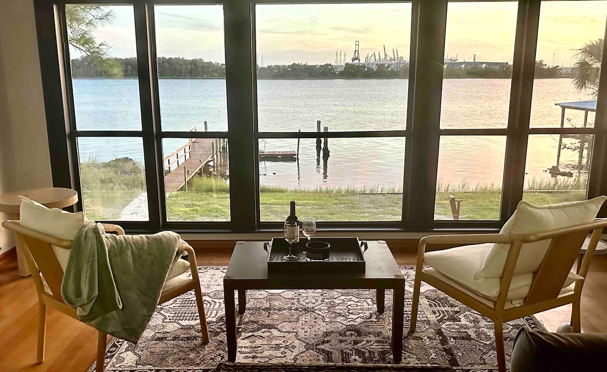 A serene seating area features two wooden chairs positioned across from a coffee table. A soft throw lies over one chair. Large windows provide an unobstructed view of the water and dock, with soft evening light illuminating the scene.