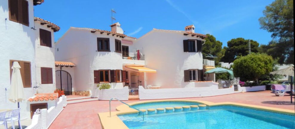 A spacious exterior view of the bungalow highlights white stucco architecture with terra-cotta roof tiles. The inviting pool is surrounded by a sun terrace, complemented by lounge chairs. Lush greenery and tall trees frame the property, under a clear blue sky.