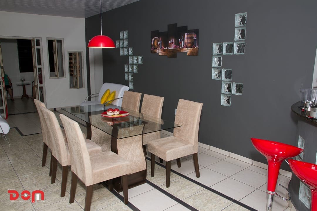 A modern dining area features a glass table surrounded by six beige upholstered chairs. A red pendant light hangs above, adding a pop of color. The walls are painted in a dark shade, complemented by vibrant wall art and glass blocks, enhancing the space's contemporary feel.