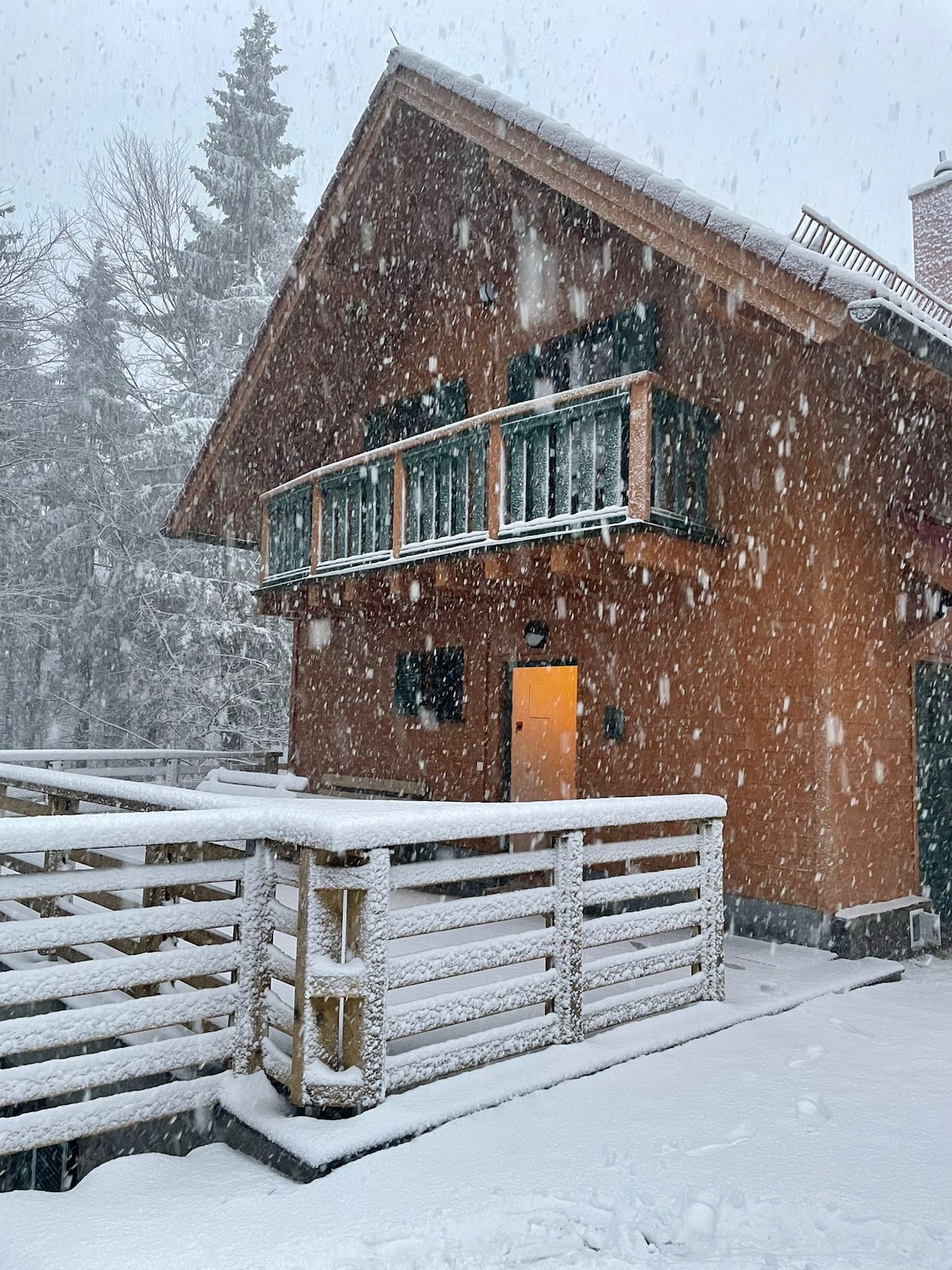 A wooden cabin is depicted covered in falling snow, with snow accumulating on the ground and surfaces. Large green shutters frame the upper balcony, which overlooks the snowy surroundings. A welcoming orange door stands out against the wooden exterior, inviting guests inside.
