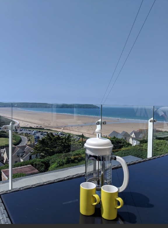 A dining table is set on a balcony, featuring two yellow cups and a French press. The scene offers an expansive view of the beach and coastline, with the clear blue sky and calm waters in the background.