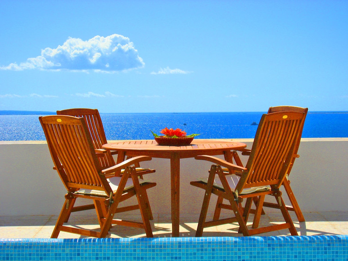 A private terrace features a wooden table surrounded by three deck chairs, offering unobstructed views of the tranquil sea and blue sky. A decorative bowl with fresh fruits is placed at the center of the table, enhancing the outdoor dining experience.