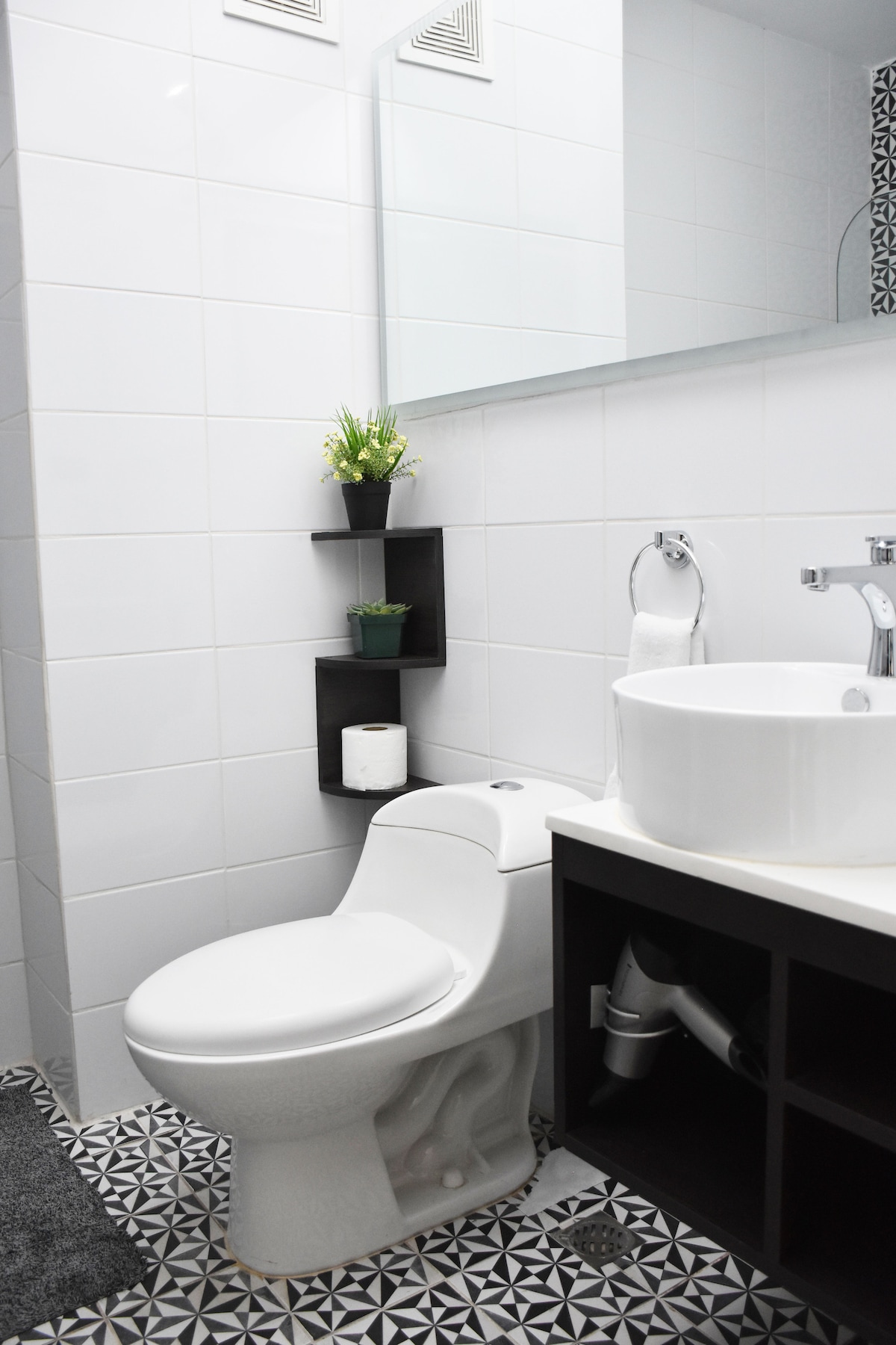 A modern bathroom is depicted, featuring a white toilet and a round vessel sink atop a dark wood vanity. A rectangular mirror is mounted above the sink. Decorative plants are placed on a wall-mounted shelf. The floor displays a geometric black-and-white tile pattern.