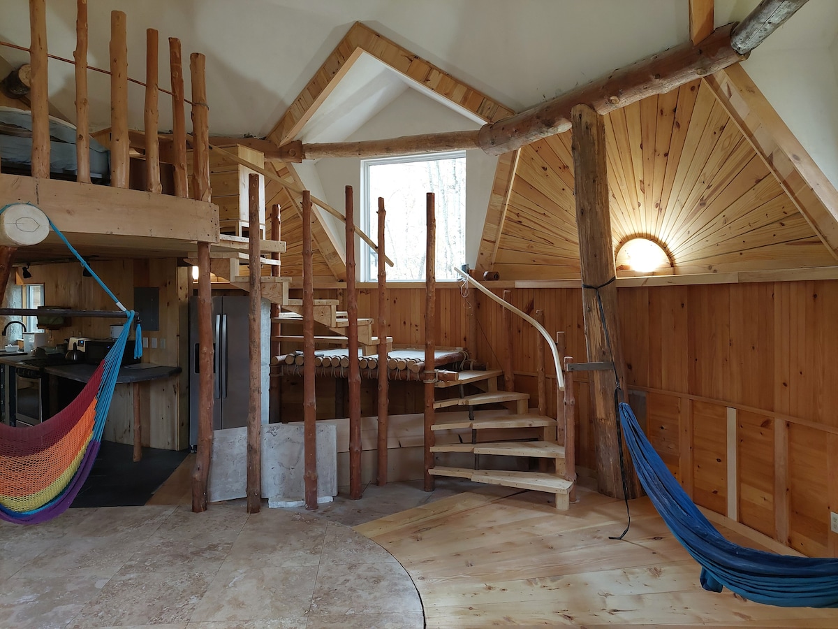 The interior features a spiral staircase with a wooden handrail, leading to a loft space. Natural wood paneling adorns the walls, complemented by a round window allowing natural light. Colorful hammocks are suspended in the corners, enhancing the cozy atmosphere.
