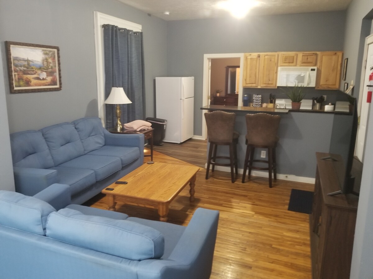 A light blue couch and loveseat are arranged in a cozy living area, complemented by a wooden coffee table. A TV stand and a lamp are positioned nearby. In the background, a dining area with barstools and a kitchenette can be seen, providing a functional space for guests.