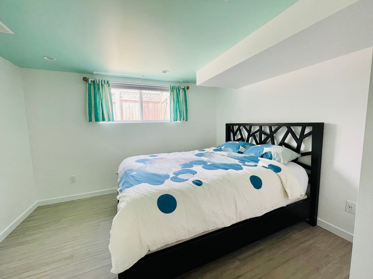 A spacious bedroom is highlighted by a queen size bed featuring a colorful duvet. Natural light fills the room through a bright window, which is dressed with turquoise curtains. The light hardwood flooring complements the modern design of the space.