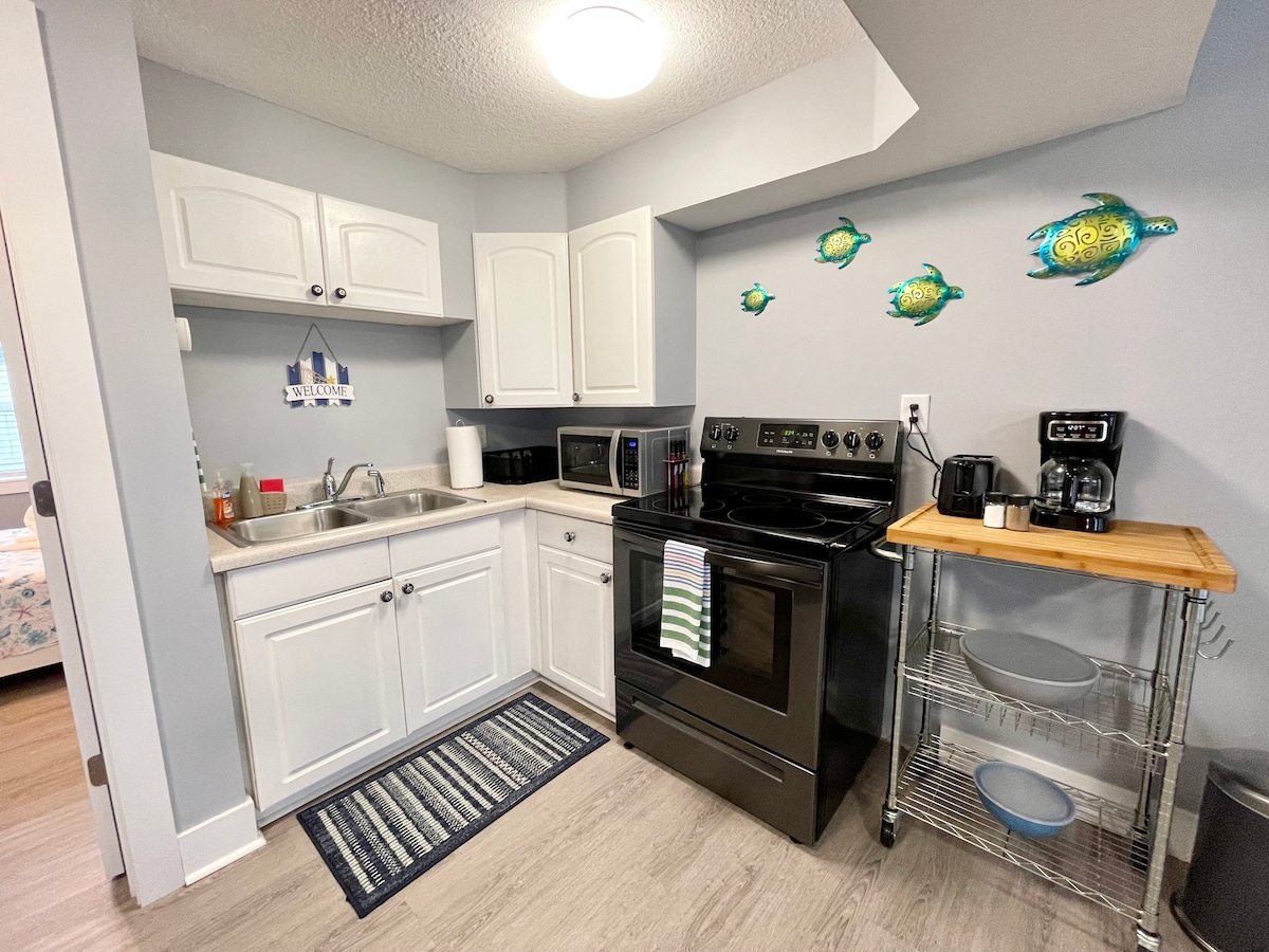 A compact kitchen space is equipped with white cabinetry and a black stove. A refrigerator and microwave are present. Kitchen utensils and a coffee maker are organized on a counter with a wooden shelf. Sea-themed decor adds a touch of character to the walls.